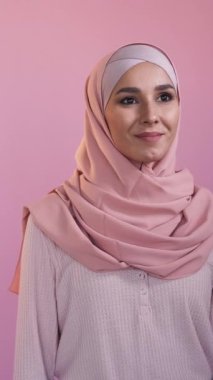 Dikey video. Mutlu kadın. İyimser bir bakış. Pastel pembeye izole edilmiş başörtüsü takmış kendinden emin, gülümseyen bir kadın yüzü..