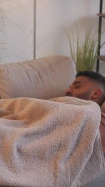 Dikey video. Evde şekerleme. Pazar dinlenmesi. Haftasonu rahatlama. Tembel bir gün. Modern oturma odasının iç kısımlarında battaniyenin altında rahat koltukta uyuyan yorgun adam..