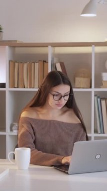 Dikey video. Evde eğitim. Bilgisayar ödevi. Akıllı bir kadın dizüstü bilgisayarda oturuyor ve modern bir fotokopi odasında kahve fincanından içiyor..