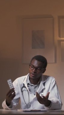 Dikey video. Eczacılık danışmanlığı. Çevrimiçi tıbbi randevu. Profesyonel tele-sağlık. Kendine güvenen erkek doktor, karanlık işyerinde boş yeri olan ilaç hapları veriyor..