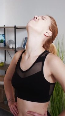 Dikey video. Boyun ağrısı. Kas ağrısı. Aşırı eğitim sorunu. Kötü duruş sorunu. Spor kıyafetli, sırt ağrısı çeken stresli bir kadın ışık odasında esneme egzersizi yapıyor..
