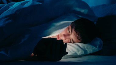 Gece, loş ışıkta yatak örtüsünün altında akıllı telefon kullanan bir adam gece yatağında uykusuzluk çektiğini gösteriyor..