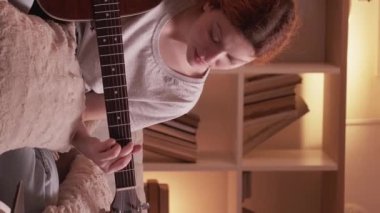 Dikey video. Çevrimiçi gitar müzik dersi. Yaratıcı bir eğlence. İlham verici müzisyen kadın telli çalgı akorları öğreniyor evdeki kanepede dizüstü bilgisayardan sanal ders izliyor..