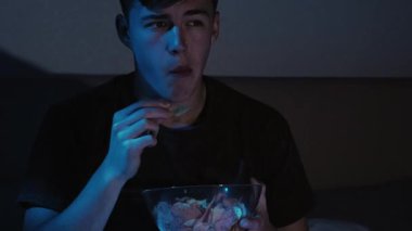 Ev sineması. Gece keyfi. Etkilenmiş adam TV 'de aksiyon filmi izlemekten zevk alıyor geç saatlere kadar yatakta oturup koyu mavi ışıklar altında cips yiyor..