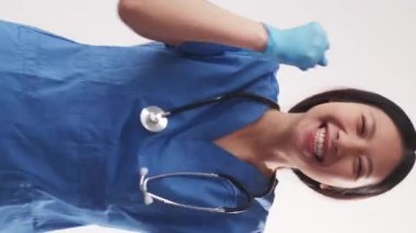 Dikey video. Heyecanlı doktor. Kazanan jest. Neşeli, mutlu hemşire kadın mavi üniforma giyiyor. Kollarını kaldırıyor. Gülümsüyor..