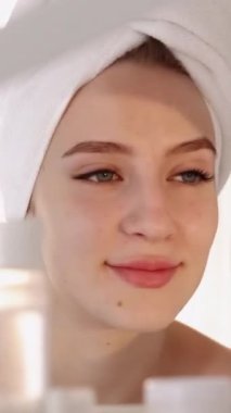 Dikey video. Yüz bakımı. Cilt tazeleme. Kozmetik tedavi. Parlak güzellik. Havluyla gülümseyen kadın spa ameliyatından sonra aynaya bakar..