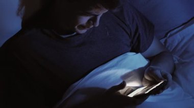 Karanlık yatak odalı adam gece yatağında uzanıyor. Akıllı telefon kullanıyor. Mavi ışıkla aydınlatılıyor..