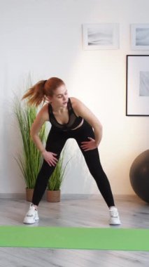 Dikey video. Evde fitness. Spor ısınması. Sabah egzersizi. Aktif yaşam tarzı. Spor kıyafetli atletik bir kadın, ışık odasının iç tarafındaki yoga minderinde kaslarını esnetiyor..