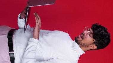 Dikey video. Çevrimiçi çatışma. İnternet kaygısı. Sanal iletişim sorunu. Kızgın, kızgın, dengesiz işadamı, kırmızı boş alana izole edilmiş dizüstü bilgisayar kullanarak yazı yazıyor..