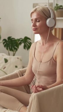 Dikey video. Ses meditasyonu. Huzurlu bir denge. Rahatlık ve rahatlık. Kulaklıklı rahat bir kadın koltukta rahat bir şekilde oturmuş evde mantra dinliyor..
