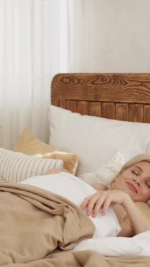 Dikey video. İyi sabahlar. Uyanma esnemesi. Gülümseyen, rahat bir kadın, uyanıp, evin içinde rahat bir yatakta oturmaktan zevk alıyor..