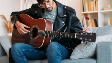 Gitar çalıyor. Yaratıcı hobi. İlham veren müzisyen akustik enstrüman tellerini akort edip evdeki kanepede ses kontrolü yapıyor..