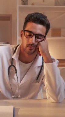 Dikey video. Yorgun doktor. Klinik aşırı çalışma. Hastane üniformalı, yorgun, profesyonel bir adam iş yerindeki çalışma masasında uyuyor..