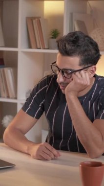 Dikey video. Yorgun bir öğrenci. Sıkıcı bir ders. İnternetten ders. Dizüstü bilgisayardan bilgi izleyen yorgun bir adam masa başında uyuyakalıyor modern aydınlık ev içinde oturuyor..