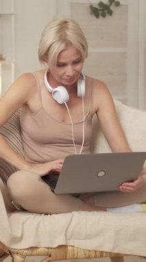 Dikey video. Serbest bir iş. Modern teknoloji. Gülümseyen kadın rahat koltukta dizüstü bilgisayarıyla oturmuş evinde, oturma odasında rahat rahat oturuyor..