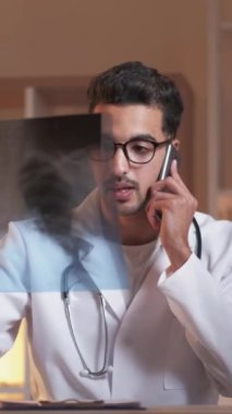 Dikey video. Telefon teşhisi. Gezici doktor konsültasyonu. Zatürree hastalığı. Akciğer röntgeni olan akciğer uzmanı akıllı telefonla konuşuyor. Kliniğin iç kısmında, çalışma masasında oturuyor..