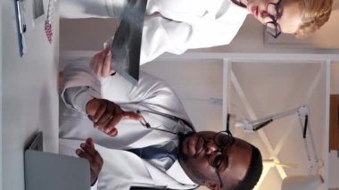 Dikey video. Klinik ekibi. Tıbbi teşhisler. Hastane sağlık hizmetleri. Erkek doktor, dizüstü bilgisayarın çalışma yerinde kadın pratisyen doktorla röntgen filmini tartışıyor..