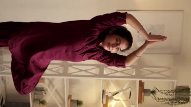 Dikey video. Yoga hobisi. Sağlıklı bir yaşam. Kapalı mekan eğlencesi. Vrikshasana ağacı pozu veren mutlu ve rahat genç bir kadın evde dikkatli hareketlerin tadını çıkarıyor..