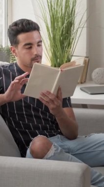 Dikey video. Sabah okuması. Ev içi rahatlık. Kitapla rahat koltukta oturan meraklı gülümseyen adam evin içinde dinleniyor..