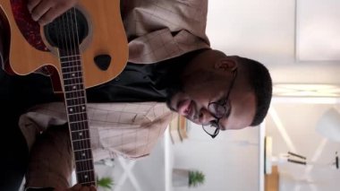 Dikey video. Şarkıcı. Erkek vokalist. Müzikal hobi. Yetenekli sanatçı evde akustik gitar çalarak şarkı söylüyor..