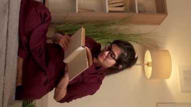 Dikey video. Kitap eğleniyor. Bilgi ve rahatlık. Evde dinleniyorum. Oturma odasında, yerde oturmuş kitap okuyan mutlu, zeki bir kadın öğrenci..