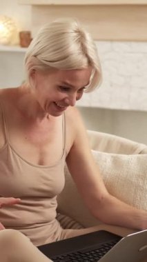 Dikey video. Sanal iletişim. Mesafe toplantısı. Çevrimiçi konferansta dizüstü bilgisayarla konuşan neşeli bir kadın evdeki rahat koltukta oturuyor..