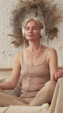 Dikey video. Sabah meditasyonu. Ses rahatla. Huzurlu bir denge. Kulaklıklı sakin bir kadın yoga pozisyonu almış, koltukta oturmuş evde mantranın keyfini çıkarıyor..