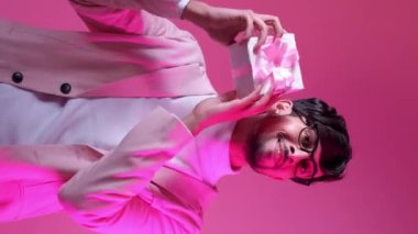 Dikey video. Romantik hediye. Sevgililer günü hediyesi. Tatil sürprizi. Neon rengi açık renk, neşeli, gülümseyen, gözlüklü, pembe kutuda izole edilmiş yakışıklı adam..
