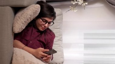 Dikey video. Telefon eğlencesi. Evde rahatla. Boş zamanlarında. Akıllı telefonlu sosyal medya uygulamasında yazan mutlu genç bir kadın oturma odasındaki rahat koltukta dinleniyor..