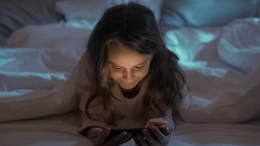 Yatma vakti. Çocuk gecesi eğlencesi. Geç saatte ev eğlencesi. Neşeli küçük kız cep telefonundan eğlenceli animasyon filmleri izlemekten zevk alıyor..