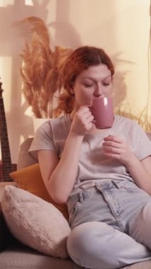 Dikey video. Afiyet olsun. Ev tatili. Sakin bir kadın çay fincanı içiyor kaygısızca dinleniyor rahat bir hayal kuruyor sıcak bir kanepe sıcak pastel renkli mobilya.