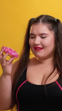 Dikey video. Tatlıyı. Sağlıksız diyet. Yüksek kalorili tatlı. Aç şişman kadın sarı arka planda izole edilmiş lezzetli pembe jöleli donut yemekten zevk alıyor..