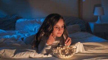 Gece filmi. Çocuk evi eğlencesi. Meraklı küçük kız TV 'de eğlenceli animasyon film izlemekten zevk alıyor patlamış mısır yiyor, gülümsüyor, karanlık bir yatak odasında mavi ışıklı bir şekilde oturuyor..