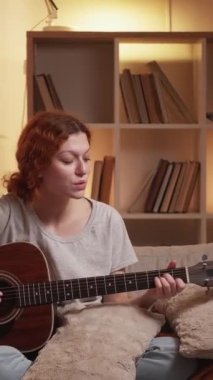 Dikey video. Gitar hobisi. Boş zamanlarında şarkı söylemek. Şarkı performansı. Kadın, evdeki kanepede akustik telle müzik çalmaktan hoşlanıyor..