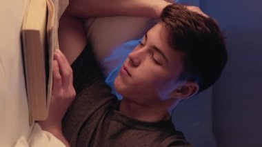 Dikey video. Yorgunluk okuyorum. Yorgun ve yorgun genç bir adam, gece yumuşak ışıkta kitap yatağıyla huzur içinde uyuyor..