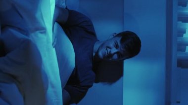Dikey video. Gece keyfi. Ev sineması. Eğlenceli bir komedi dizisi izlemekten zevk alan neşeli bir adam. Koyu mavi bir ışıkta geç saatte yatakta gülüyor..
