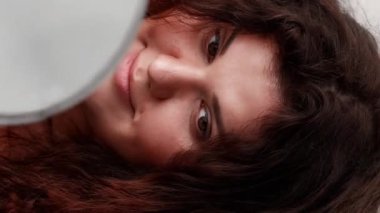 Dikey video. Güzellik bakımı. Sağlıklı saçlar. Sabah rutini. Kıvırcık bukleli kilitlere dokunan şişman, güler yüzlü kadın aynaya bakıyor..