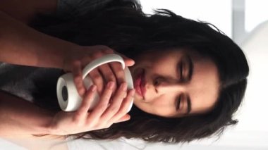 Dikey video. Kahve gevşedi. Ev istirahatı. Hafta sonu tatili. Tatmin olmuş bir kadın bir fincan latte içiyor güneşli bir sabah güneşinin tadını çıkarıyor..