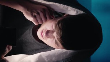 Dikey video. Karanlık odada battaniyenin altında akıllı telefon kullanan genç. Geceleri parlayan yüzü olan..