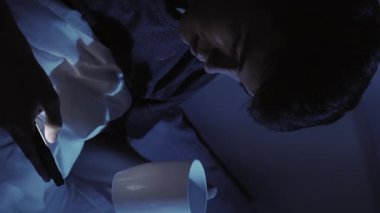 Dikey video. Karanlık odada uyuyan adam yatakta akıllı telefon fincanı kullanmadan önce rahatlama bağlantısı kurar ve rahat bir gece rutini yaratır..