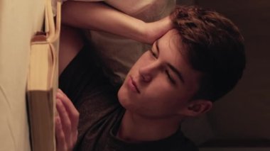 Dikey video. Gece keyfi. Huzurlu adam yatakta kitap okumaktan zevk alıyor. Yatak odasında huzurlu bir ortam yaratıyor..