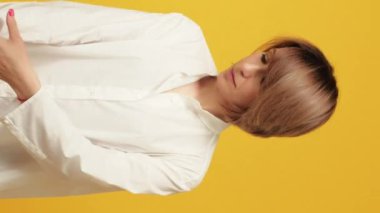 Dikey video. Kıyafet deneyen alışveriş yapan kadın. Beyaz resmi bluz giyen olgun bir kadın manken sarı üzerine izole edilmiş kıyafet seçiyor..