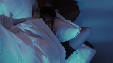Dikey video. Gece uykusuzluğu. Uyku sorunu. Huzursuz adam yatakta dönüp başını yastıkla örtüyor loş mavi ışığın altında.
