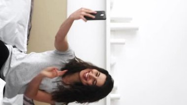 Dikey video. Selfie eğlencesi. Milenyum kadını. Çevrimiçi blog yazarı. Akıllı telefondan fotoğraf çeken mutlu bir kadın poz veriyor. Yumuşak yatak aydınlığında rahat bir yatak odasında oturuyor..