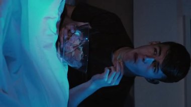 Dikey video. Ev sineması. Gece keyfi. Etkilenmiş adam TV 'de aksiyon filmi izlemekten zevk alıyor geç saatlere kadar yatakta oturup koyu mavi ışıklar altında cips yiyor..