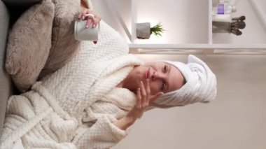 Dikey video. Güzellik tedavisi. Stres atmak. Rahatlamış, dingin, genç bir kadın banyo havlusu ve duştan sonra sıcak çay içen yumuşak bir bornoz..