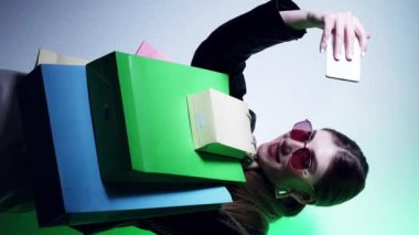 Dikey video. Alışveriş selfie 'si. Kara Cuma. Alışverişkolik yaşam tarzı. Gözlüklü göz kamaştırıcı kadın telefon kamerasında yeşil ışık altında poz veriyor..