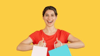 İyi alışverişler. Kırmızı çantalı mutlu bir kadın sarı boş bir alanda izole edilmiş heyecanını gösteren bir jest gibi başparmak kaldırıyor..
