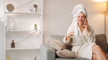 Kız kıza. Ev spası. Temiz tedavi. Vücut sağlığı. Beyaz banyo havlusu içinde rahat bir şekilde gülen genç kadın telefonda konuşan, duştan sonra sıcak içecek içen yumuşak bornoz.