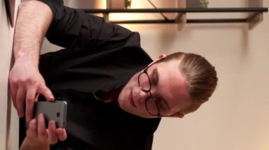 Dikey video. Kariyer gelişimi. Cep telefonu çalışıyor. Gözlüklü, siyah tişörtlü, akıllı telefonlu başarılı genç adam internette cihazın üzerinde oturuyor..